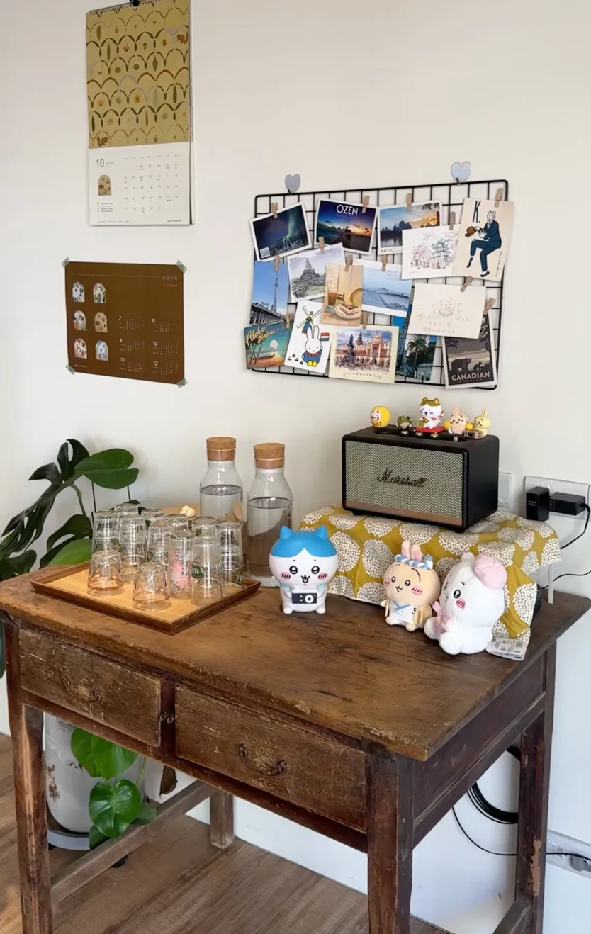 日知常屋的自助水杯區,老木桌上擺著玻璃杯與水瓶,牆面掛著明信片與月曆,角落擺放音響與公仔
